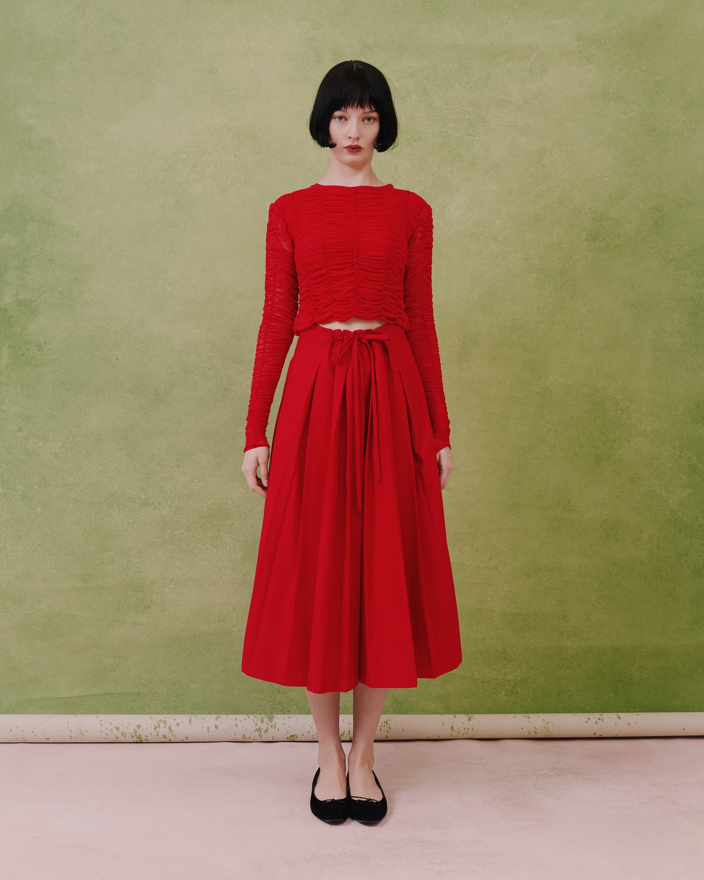 Model wears the Amelia Toro ruched tulle long-sleeve crop top in red with a pleated midi skirt, front view